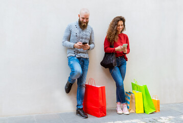 happy couple of a bearded bolt man and a curly caucasian woman standing against a wall background with shopping bags in the city center during black friday sales. girl and boy interacting with phones