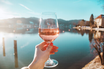 Female hand with glass of rose wine. Cozy pier on the coast of the lake Tegernsee. Alps mountains in Bavaria. Beautiful landscape with sun rays in Germany. Adventure in Europe, travel photo.