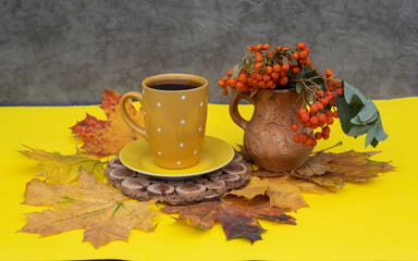 Wall Mural - There is a cup of tea and yellow leaves on a yellow background, not on a stand. Nearby is a clay jug with a bouquet of rowan berries.