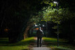Young adult alone man in warm clothes walking on sidewalk through alley of trees under lamp light in autumn night. Spending time alone in nature. Peaceful atmosphere. Back view.