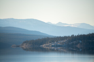 hazy mountain and lake landscape with calm, reflection shore and wilderness woods with spruce and pi