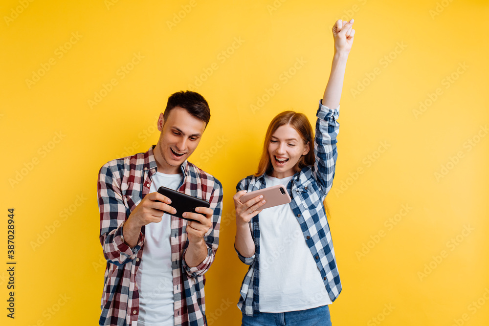 happy couple, a man and a woman, play video games on a smartphone, rejoice and show a winning gesture, on an isolated yellow background - obrazy, fototapety, plakaty 