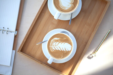 Top view two cups of coffee latte art foam is holding by woman hand with unfocused placed eyeglasses and notepad on wooden table.