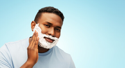 Wall Mural - grooming and people concept - african american man applying shaving cream to his face over blue background