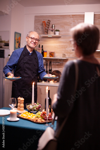 Senior man serving wife while celebrating their relationship with tasty and and wine. Elderly old couple talking, sitting at the table in kitchen, enjoying the meal, celebrating their anniversary .
