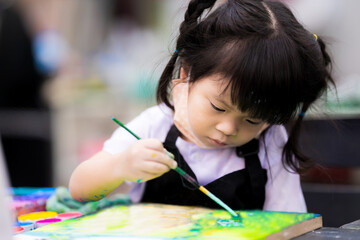 Cute girl is coloring on canvas. Child catch brushes and learn to painting water color. Children wear a black waterproof apron. Adorable little  kid is 3 years old.