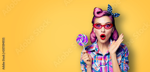 Purple head excited very surprised woman with lollipop. Pinup girl in rose pink red glasses with wide opened mouth, eyes. Retro and vintage concept. Yellow orange color background with text copy space © vgstudio