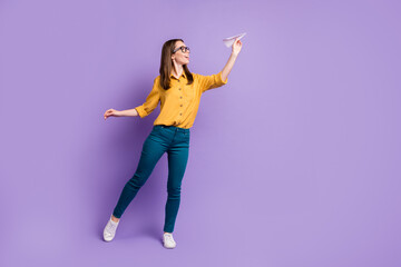 Poster - Full length profile photo of girl hold paper plane empty space wear specs yellow shirt blue trousers sneakers isolated violet color background