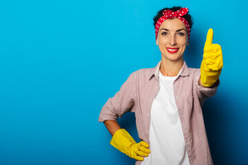 Smiling young woman in cleaning gloves showing thumb up gesture, class gesture on blue background.