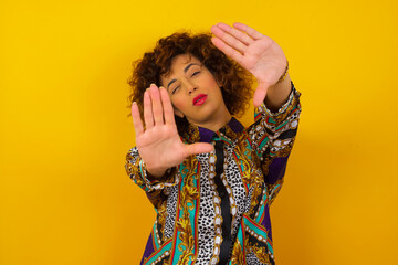 Wall Mural - Portrait of smiling beautiful young woman standing against yellow background wearing casual clothes, looking at camera and gesturing finger frame or stop sign with both hands.