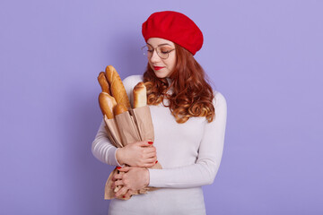 Portrait of beautiful young woman holding paper bag with bread on lilac background, girl with fresh fragrant long loaf, wearing white casual shirt and red beret, has wavy hair.