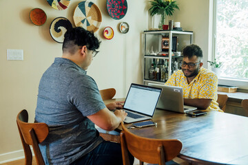 Wall Mural - Gay couple working from home together on laptop computers sitting at dining room table 