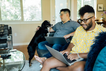 Wall Mural - happy gay couple working from home with laptops on couch with their australian shepherd