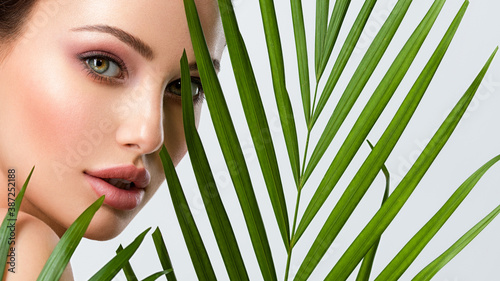 Young beautiful woman with healthy skin of face and palm leaves. Closeup fresh face of an attractive caucasian girl with green plants. Model with bright brown eye makeup. Skin care concept.