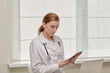 © Evgenia - Portrait of a young woman doctor in a white medical coat with a tablet in her hands. White background.