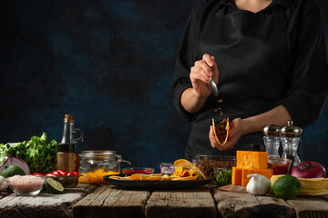 Wall Mural - Professional chef in black uniform puts the filling on corn tortilla for cooking tacos on dark blue background. Traditional mexican cuisine. Concept of tasty street food. Frozen motion.