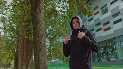 Portrait of a hipster young man putting on a hood while looking at the camera. Close up view of millennial boy in dark hoodie smiling standing on the street. People emotion concept