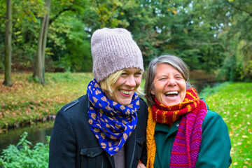 two women in their 50s hugging each other and smiling in park in autumn