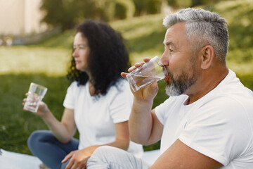 Senior couple is doing yoga outdoors. Stretching in park during sunrise. Brunette in a white t-shirt.