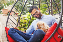 Handsome Indian Man Sitting On The Hanging Chair In The Garden With His Cute Little Dog