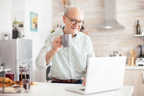 Cheerful senior man during video conference in kitchen while enjoying breakfast and cup of coffee. Elderly person using internet online chat technology video webcam making a video call connection