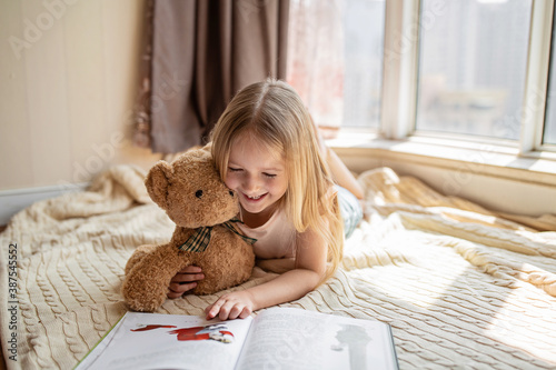 Fotografía Cute little caucasian girl in casual clothes reading a book with stuffed teddy bear toy and smiling while lying on a floor near window in the room