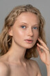© DimaBerlin - Closeup of pretty young woman face with blue eyes, curly natural blonde hair, has no makeup, touching her soft skin, standing shirtless with bare shoulders, looking at camera. Studio grey background