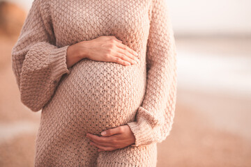 Pregnant woman wearing cozy knitted sweater holding tummy with hands outdoors close up. Motherhood. Maternity. Heathy lifestyle.