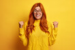 © WHstudio Leushin N - Euphoric overjoyed redhead woman raises clenched fists makes yes gesture excited by excellent news wears spectacles and yellow jumper celebrates getting prize stands indoor. Success victory concept