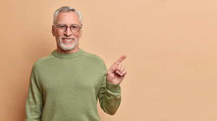 Smiling positive senior man with grey hair and beard points right and shows perfect copy space has brilliant smile white teeth wears optical glasses casual jumper poses against beige background