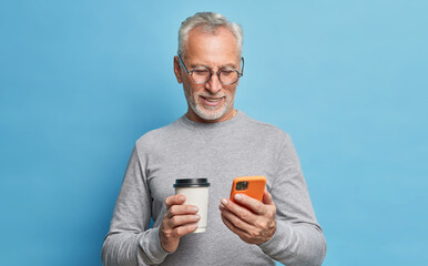 Cheerful modern senior man uses cellphone for communication types text message on phone screen holds paper cup of coffee scrolls internet pages dressed in casual clothes isolated over blue wall