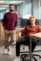 Poster - Two young serious businessmen in casualwear sitting and standing by workplace