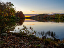 Sunset Over Keystone Lake In West Moreland Country In The Laurel Highlands Of Pennsylvania In The Fall With The Fallen Leaves In The Foreground, The Colorful Orange Sky In The Background And Trees.