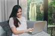 © skarie - Pretty smiling young woman sitting on sofa and working on laptop