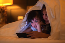 Asian Girl Children Enjoy Leisure Time With Mom Relax In Bedroom Using Smartphone At Night, Watch Video About Teaching Content While Lying In Bed Under The Blanket Together.