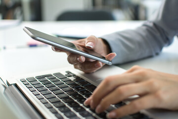 close up of female hands holding smart phone and typing on laptop keyboard.