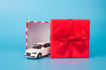 Photo of toy little white modern fashionable car driving out of present box with big red bright bow isolated over blue background