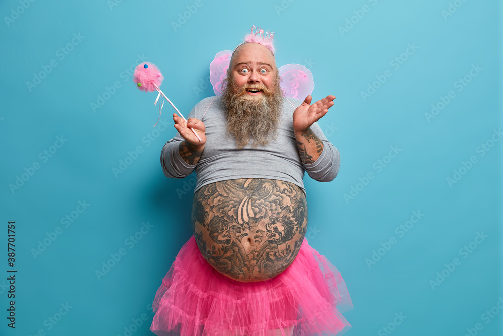 Funny happy bearded man has image of fairy holds magic wand poses with big tattooed belly over blue wall entertains children on party poses against blue background. Adult male dressed like princess - obrazy, fototapety, plakaty 