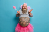 Fototapeta  - Funny happy bearded man has image of fairy holds magic wand poses with big tattooed belly over blue wall entertains children on party poses against blue background. Adult male dressed like princess
