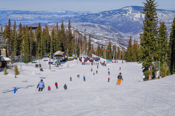 View of Colorado ski resort in winter: people skiing and snowboarding to base of chairlift; forest and mountains in background