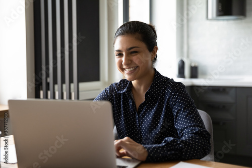 Smiling young Indian female worker sit at desk in office look at laptop screen working distant. Happy millennial ethnic woman employee consult client online on computer, typing texting on gadget.