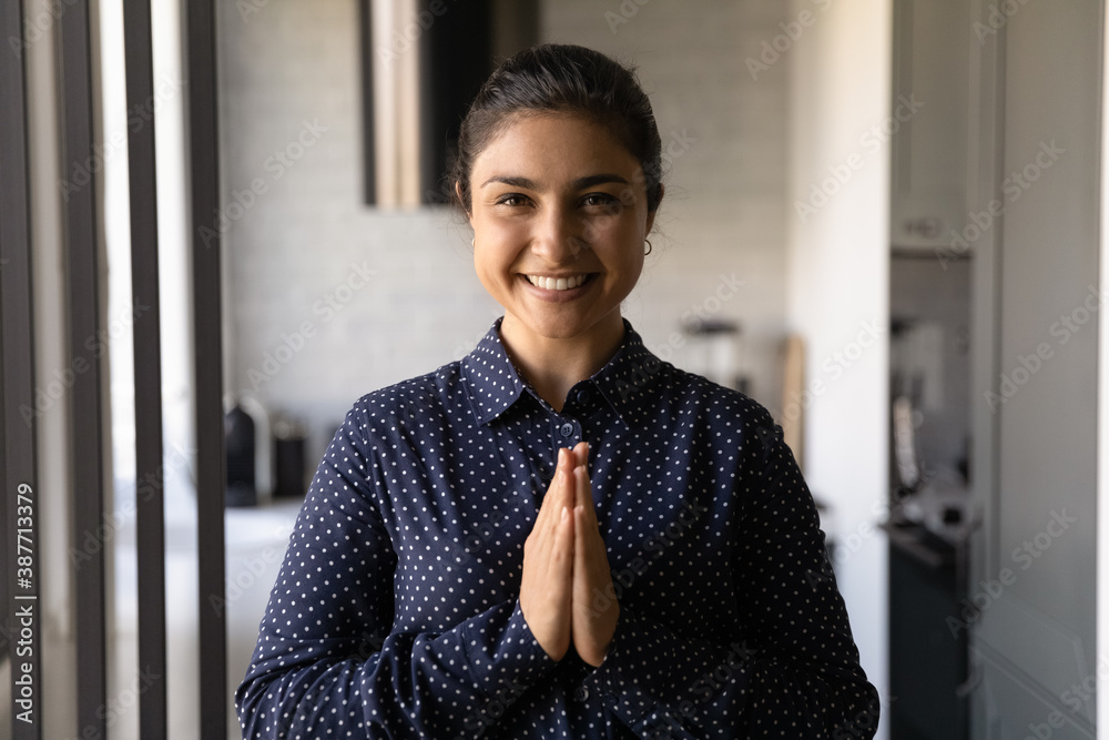 Portrait of smiling young Indian woman look at camera pose hold hands in Namaste gesture. Happy millennial ethnic female feel religious superstitious praying at home. Religion, faith concept. - obrazy, fototapety, plakaty 