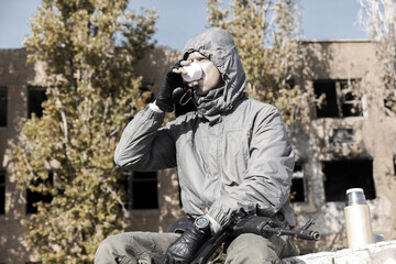 Wall Mural - a confident male stalker in a jacket with a hood sits and drinks tea from a thermos, weapons on his lap, against the background of a ruined house