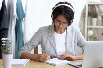 Wall Mural - Focused millennial Caucasian female in headphones make notes study distant on computer sitting at desk. Young woman in earphones write take online course or training on laptop. Education concept.