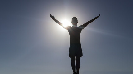 silhouette of a man with open arms raised in an attitude of triumph, from behind you can see the brightness of the sun, the sky is clear and blue