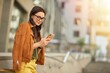 Side view of a young beautiful business woman using her smartphone, texting sms or chatting with friend while standing against blurred urban background outdoors