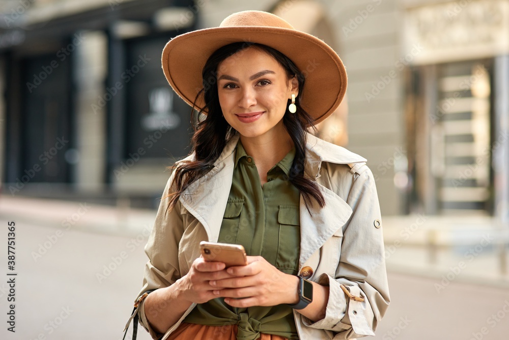 Portrait of a young beautiful stylish woman wearing autumn coat and hat holding her smartphone, looking at camera and smiling while standing on city street - obrazy, fototapety, plakaty 