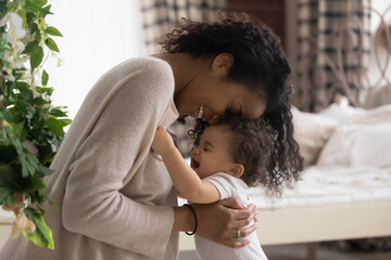 Wall Mural - Happy loving young African American mother hug embrace paly with little cute ethnic baby infant at home. Smiling caring biracial mom feel playful enjoy tender family moment with small toddler child.