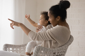 Wall Mural - Loving young African American mother cuddle in chair at home with little baby child playing together. Caring happy biracial mom hug embrace have fun with small newborn daughter. Motherhood daughter.