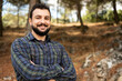 Man with beard and plaid shirt of aesthetic lumberjack crosses his arms in the middle of nature in portrait looking at camera